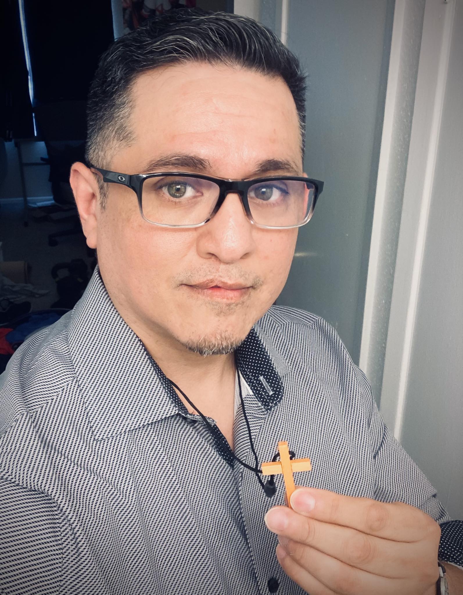 Man with dress shirt on holding a  wooden cross hung on his neck by a black string. He is wearing glasses.