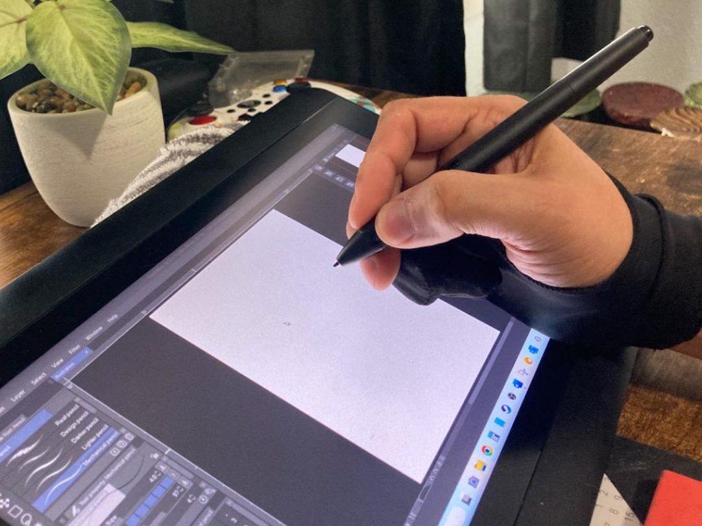 hand hovering over drawing tablet on a wooden table next to a small plant.